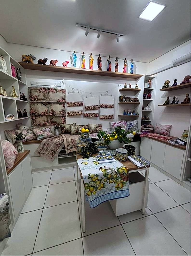 Sala do Bazar com almofadas floridas, toalhas bordadas e imagens em cerâmica expostas em prateleiras e sobre a mesa