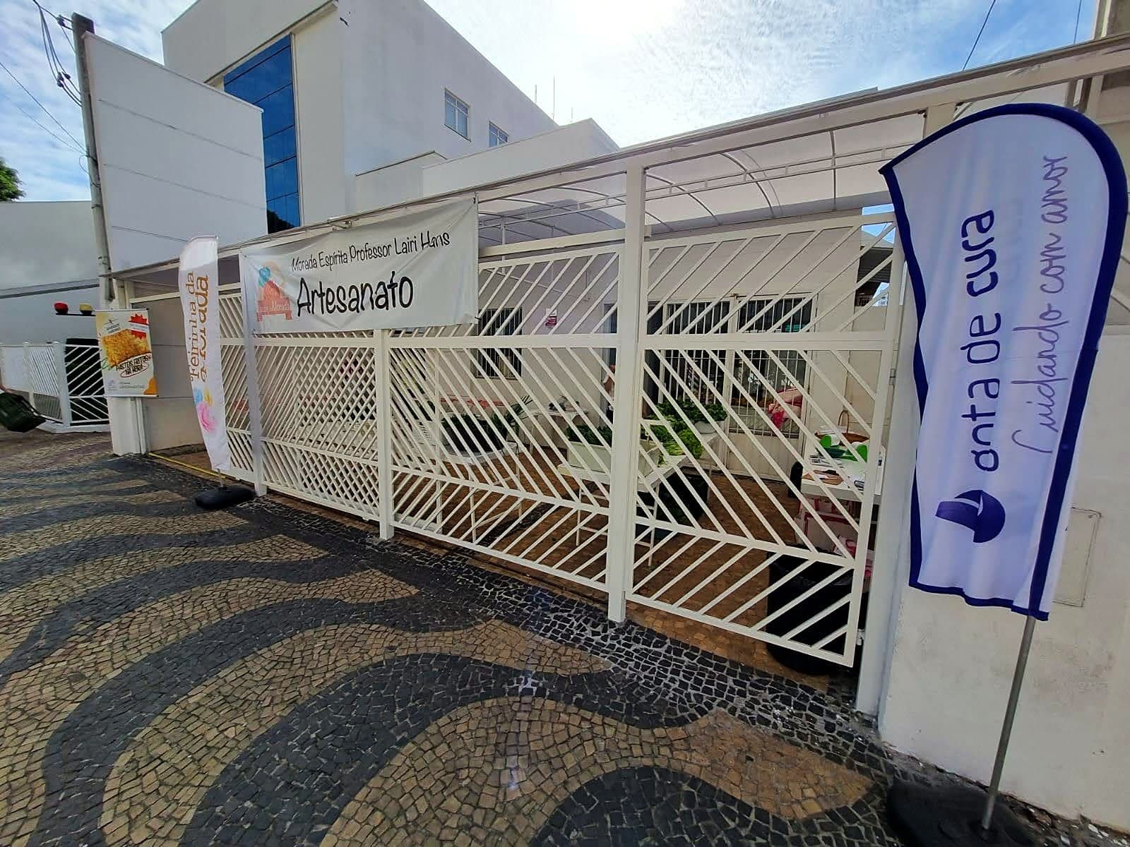 Entrada da Loja da Morada, com as bandeiras da Feirinha, do Artesanato e da Gota de Cura, na calçada de mosaico da Rua José Paulino