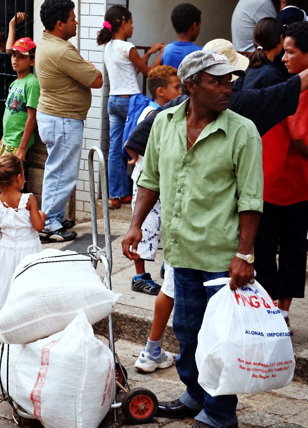 Homem com uma sacola de alimentos na calçada, ao lado de um carrinho com sacos grandes, entre crianças e adultos
