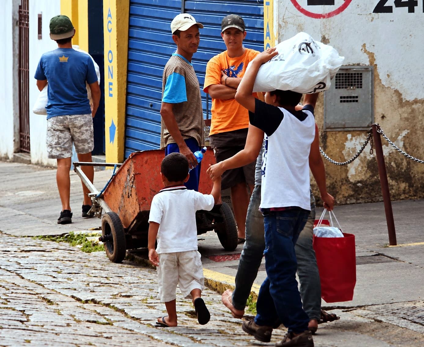 Crianças e adultos carregando sacolas de alimentos numa rua de paralelepípedos, ao lado de um carrinho de mão