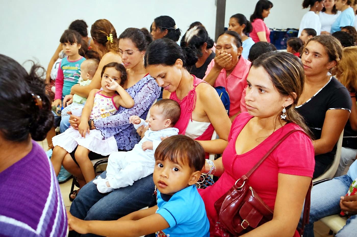 Mães com bebês e crianças no colo, sentadas lado a lado numa sala, aguardando atendimento