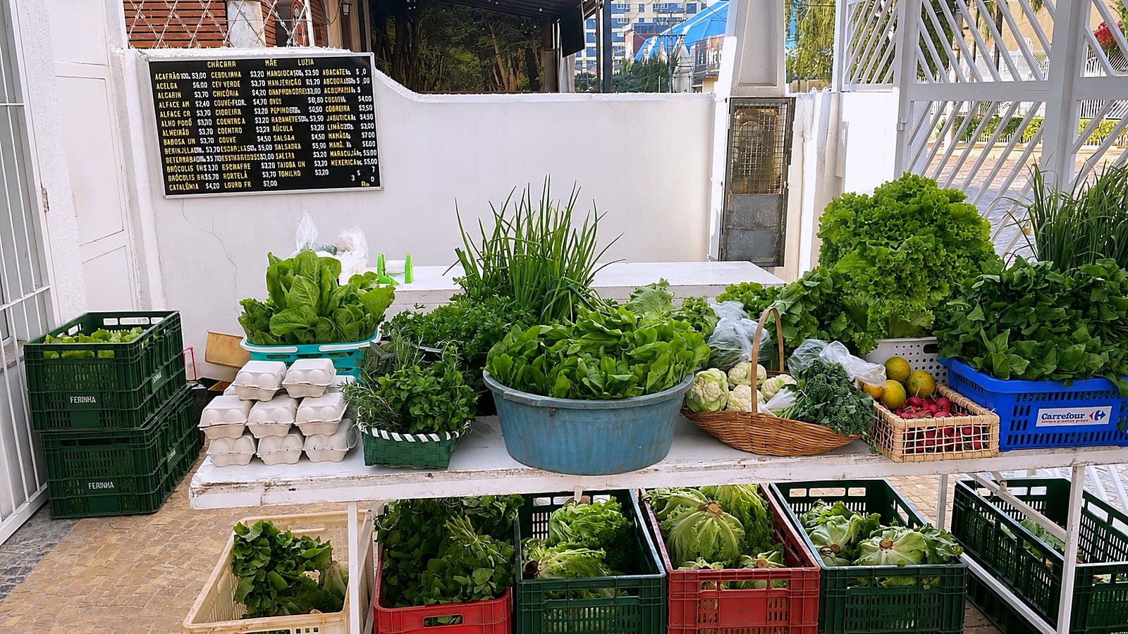 Banca da feira com alface, cebolinha, couve-flor, ovos e temperos frescos, e o quadro de preços ao fundo