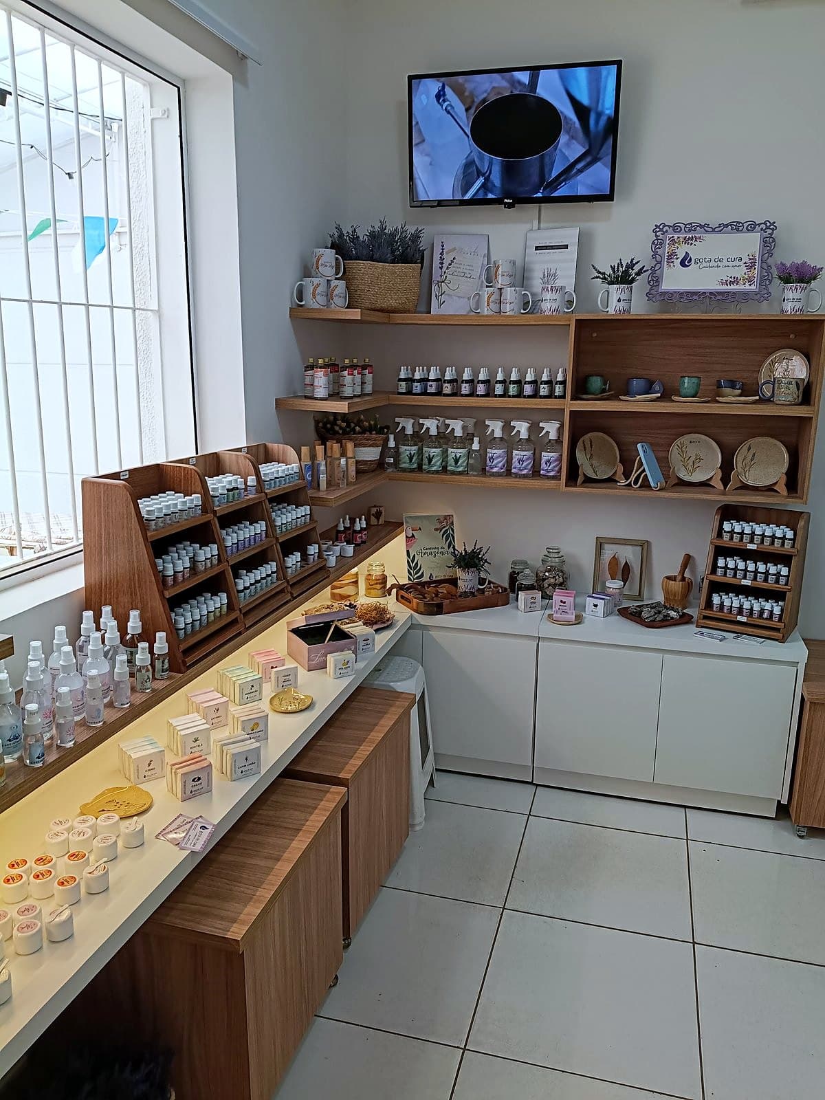 Prateleiras da Gota de Cura com óleos essenciais, sabonetes e outros produtos de aromaterapia, ao lado de uma janela