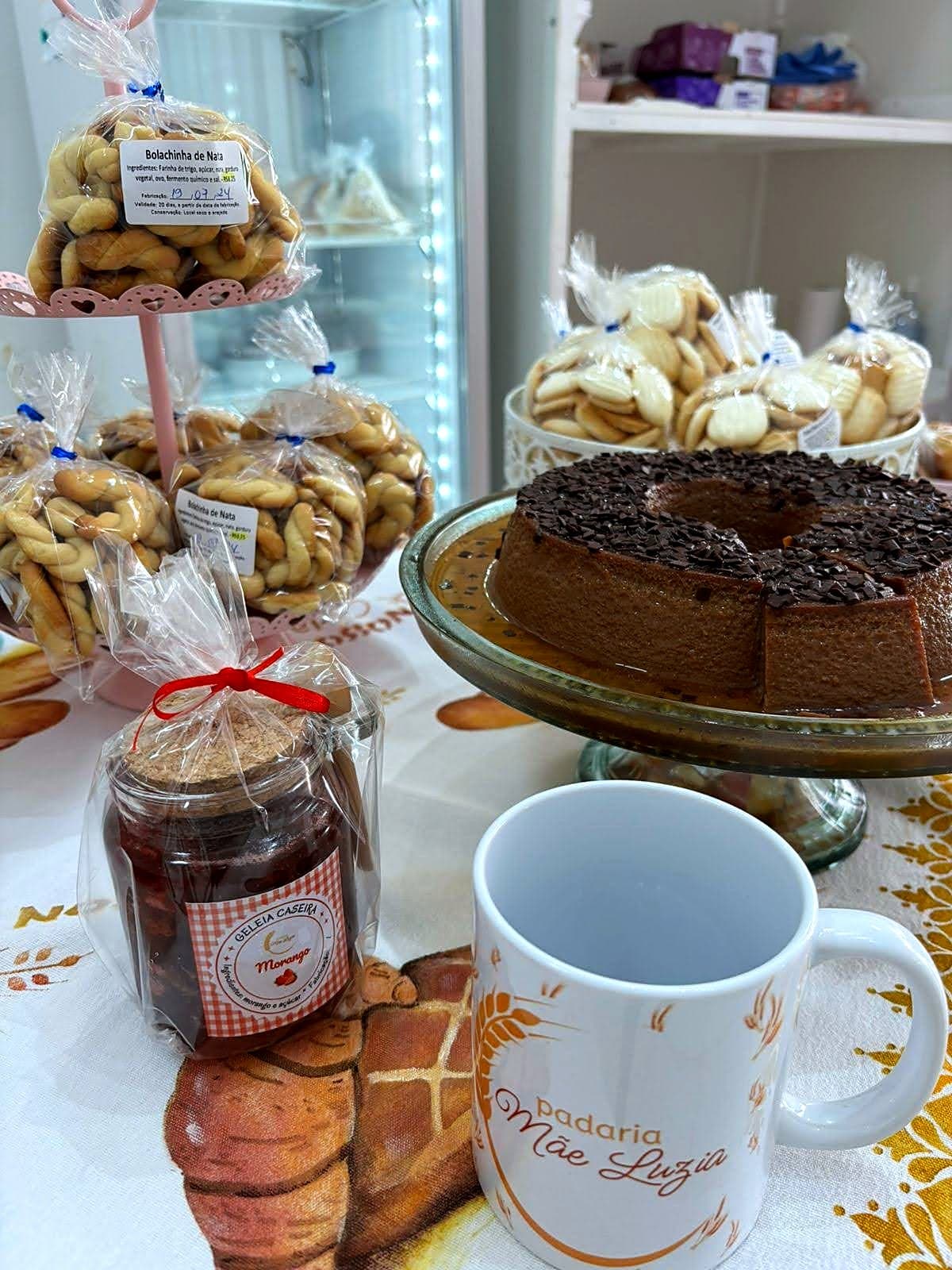 Bolo de chocolate, biscoitos amarrados em saquinhos e geleia de morango caseira sobre a mesa, ao lado de uma caneca da Padaria Mãe Luzia